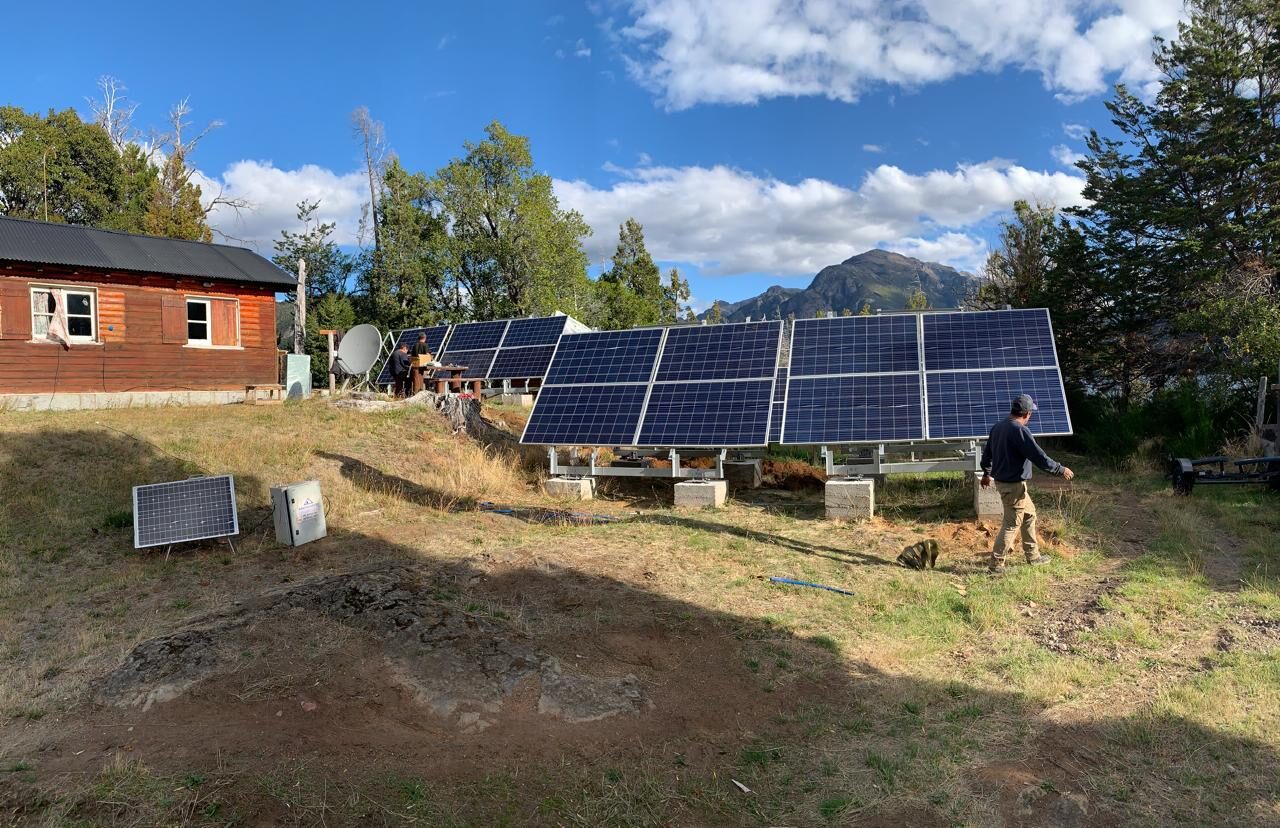 Un conjunto de paneles solares instalado sobre el pasto, junto a una casa de madera, con la montaña al fondo